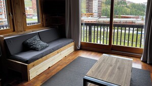 A modern room with a wooden floor, a grey sofa, a wooden table, and a large window with a view of buildings and greenery. - Châlet located in PLAGNE Centre, rated 4 Paradiski crystals and furnished for tourism, (La Plagne Tarentaise)