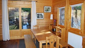 A wooden cabin interior with a dining table, benches, a small kitchen area, and large windows with curtains. - Châlet located in PLAGNE Centre, rated 4 Paradiski crystals and furnished for tourism, (La Plagne Tarentaise)