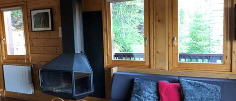 Een gezellig chaletinterieur met houten details, een open haard, een bank met kussens en ramen met uitzicht op een bos.
