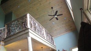 Une pièce aux hauts plafonds, avec un plafond en bois, un balcon doté d’une balustrade ouvragée et un ventilateur de plafond.