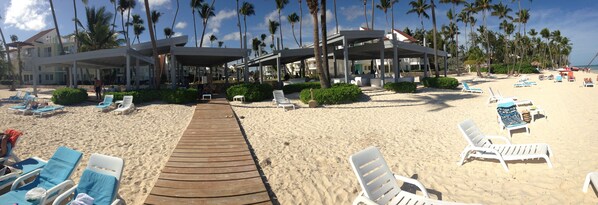 A beach with a wooden walkway leading to a covered area with lounge chairs and umbrellas.
