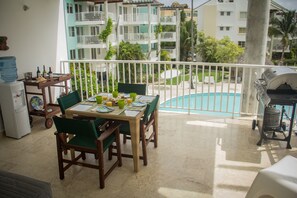 Een balkon met een tafel, stoelen, een barbecue en een zwembad op de achtergrond.