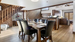 A dining area with a wooden table set for six, surrounded by leather chairs. A staircase with wooden railings leads to an upper level.