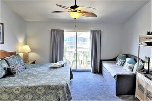 A hotel room with a bed, a sofa, a ceiling fan, a window with curtains, and a view of buildings outside.