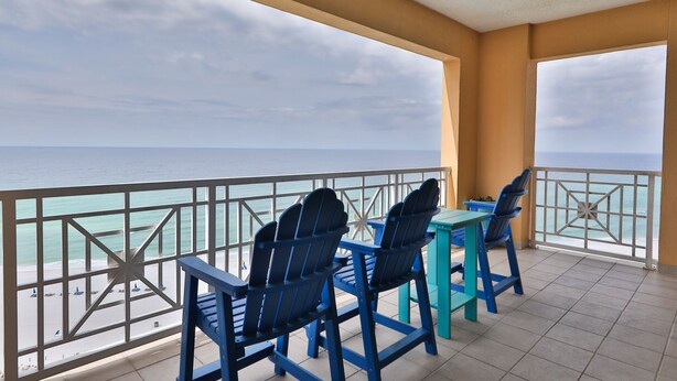 Un balcón con dos sillas Adirondack azules y una mesa turquesa, con vista a una playa y una perspectiva despejada del océano.