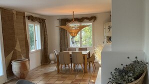 Ein Essbereich mit Tisch und Stühlen, einer hängenden Leuchte, einem großen Fenster mit Vorhängen und einer Topfpflanze.