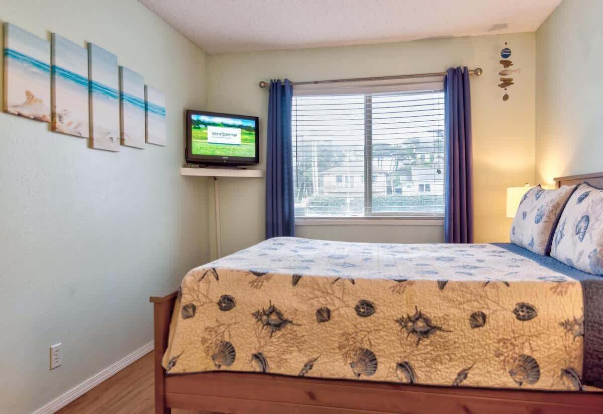 A bedroom with a bed, a television on a stand, a window with curtains, and a wall with framed pictures.