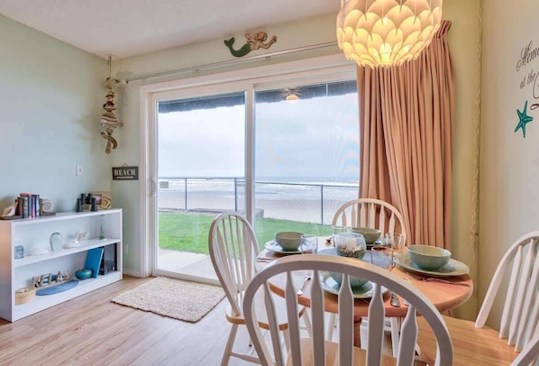 Enjoy meals in this dining area with a table for four, a shelving unit, and a sliding glass door leading to a balcony with a sea view.