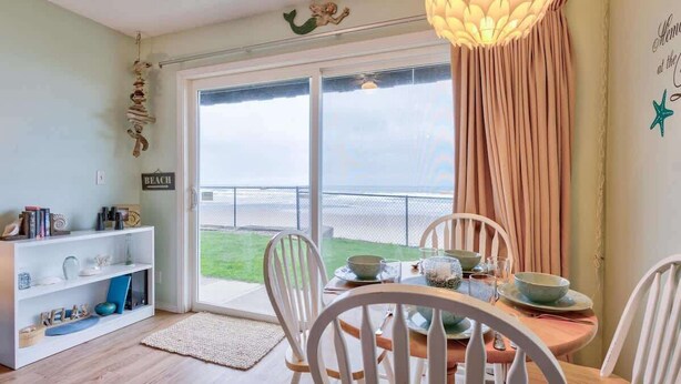 Een eetgedeelte met een tafel voor vier personen, witte stoelen, een witte boekenkast en een groot raam met uitzicht op het strand.