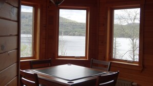Una cabaña de madera con una mesa y sillas, ventanas amplias que dan a vistas de un lago y un bosque.