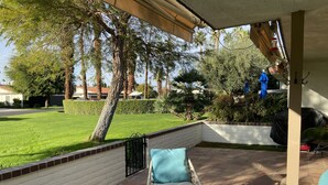 Een overdekt terras met tuinmeubilair, een bakstenen muur en uitzicht op een verzorgd gazon en bomen.