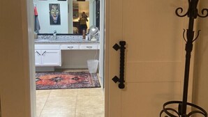 Een keuken met witte kastjes, een zwart aanrecht en een vloerkleed in rood en zwart. Er staat een zwart metalen rek aan de rechterkant van de deur.
