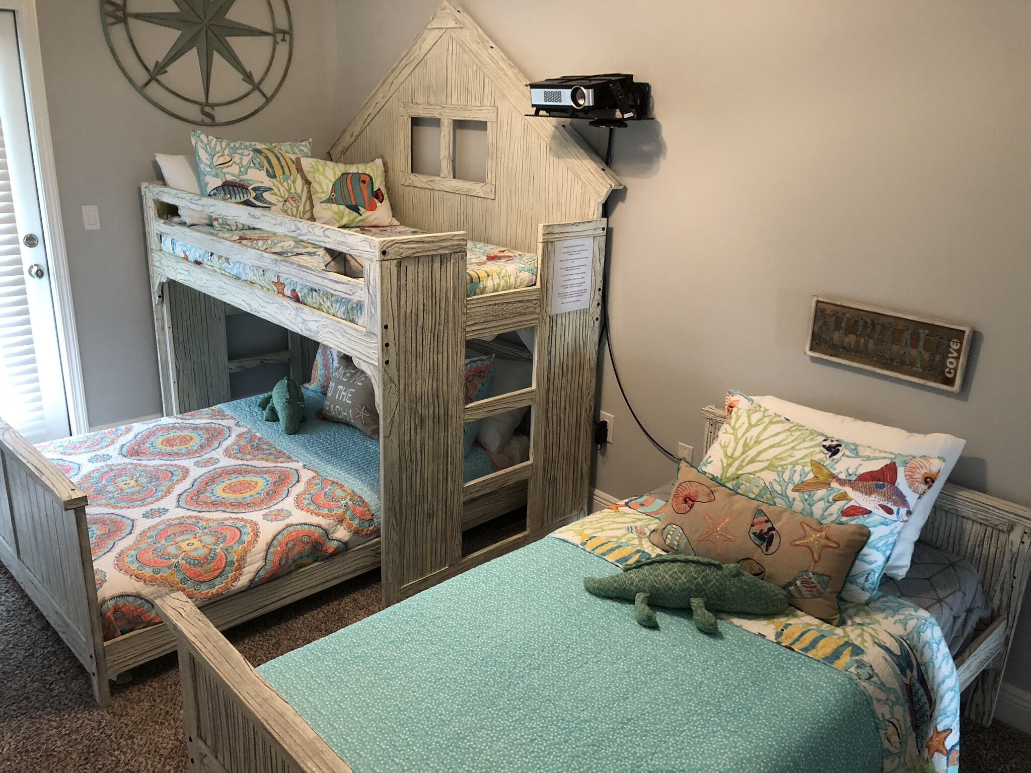 A bunk bed room with a wooden house design, a projector mounted on the wall, and a nautical-themed bedspread.