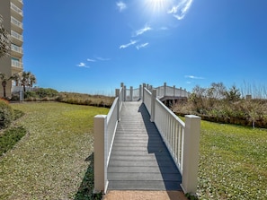 A wooden boardwalk leading to a white bridge, surrounded by grass and vegetation.