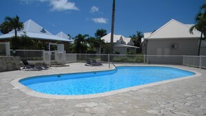 A swimming pool with stone tiles, lounge chairs, and a fenced area with palm trees. - Cause desistement Magnifique duplex jardin privatif, piscine et si dispo clio3 (Saint-François)