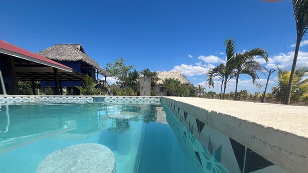 A clear swimming pool with a tiled edge, a thatched-roof structure, and palm trees in the background. - Surf, Sun & Serenity – 2BR Beachfront Home at Playa Tesoro (Between Salinas Grandes and Puerto Sandino)