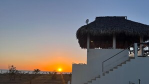 A building with a thatched roof and a staircase leading to a balcony overlooking a beach at sunset. - Surf, Sun & Serenity – 2BR Beachfront Home at Playa Tesoro (Between Salinas Grandes and Puerto Sandino)