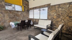 Una habitación con pared de piedra, un sofá blanco, una mesa de comedor con sillas y una ventana con celosía de metal.