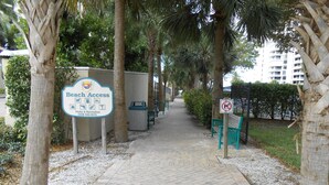 Un sentier menant à un panneau indiquant l’accès à la plage, bordé d’arbres et agrémenté d’un banc.