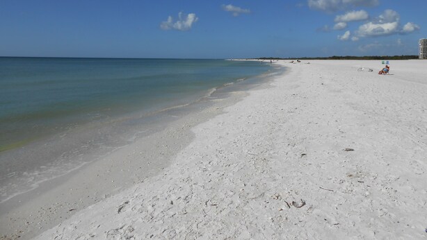 Une vaste plage de sable fin, avec une eau d’un bleu limpide et quelques personnes visibles au loin.