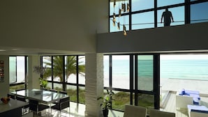 A modern interior with large windows overlooking a beach, a dining area, and a seating area with a view. - Pure luxury at #Sunset Wave on the Beach (Treasure Island)