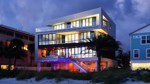 A modern beachfront building with a prominent staircase leading to the entrance, surrounded by sand and vegetation. - Pure luxury at #Sunset Wave on the Beach (Treasure Island)