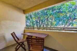 Ein Balkon mit Holztisch und -stühlen, Blick auf grüne Bepflanzung durch ein Fenster.