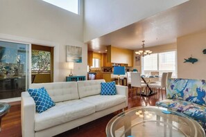 A living room with a sofa, a glass coffee table, and a colorful armchair. There is a dining area with a table and chairs, and a kitchen in the background.