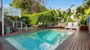Une piscine dotée d’une terrasse en bois, entourée d’une végétation luxuriante et accessible par un escalier menant à un niveau supérieur.