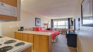 Ein kompaktes Hotelzimmer mit Küchenzeile, einem Bett mit roter Bettwäsche, einem Schreibtisch und Blick auf den Ozean.
