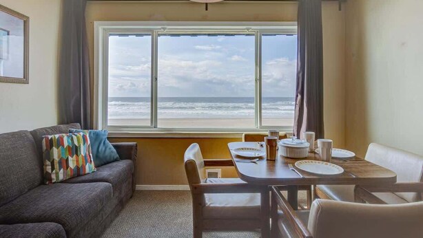 Ein Zimmer mit großem Fenster und Meerblick, einem gedeckten Esstisch sowie einer Couch mit farbigem Kissen.