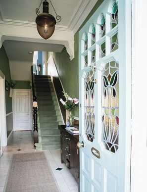 Un couloir avec un escalier, une porte décorative ornée de vitraux et un luminaire suspendu.