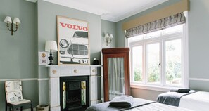 Une chambre avec deux lits simples, une cheminée et une grande fenêtre avec des rideaux.
