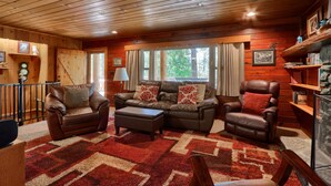 A cozy cabin interior with a wooden ceiling, leather furniture, and a large window offering a view of trees.