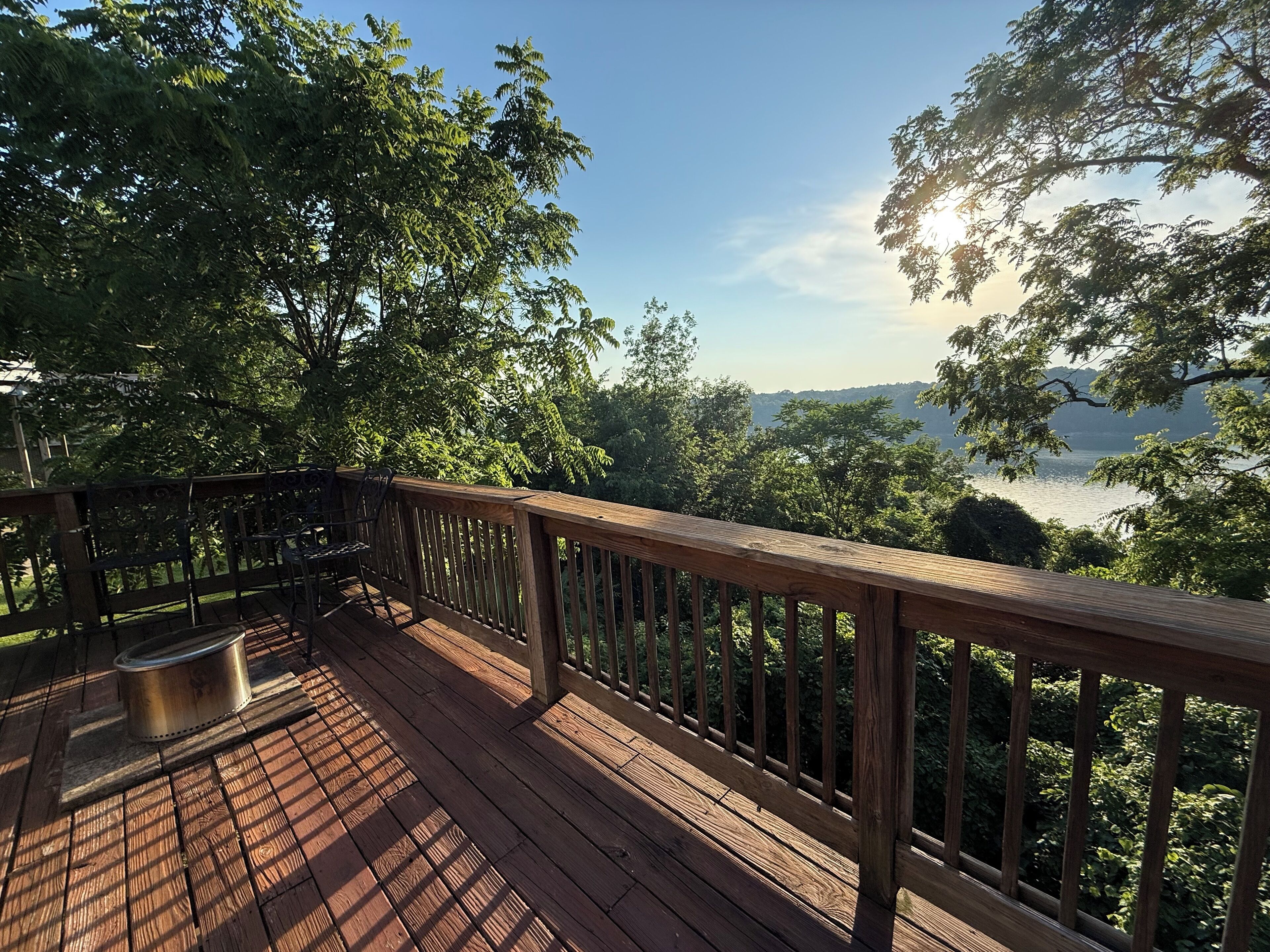 Una terraza de madera con una parrilla de metal, con vistas a un lago rodeado de árboles y colinas.