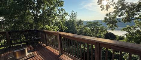 Una terraza de madera con una parrilla de metal, con vistas a un lago rodeado de árboles y colinas.