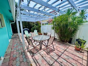 A covered patio area with a round table and chairs, surrounded by potted plants and a white fence.
