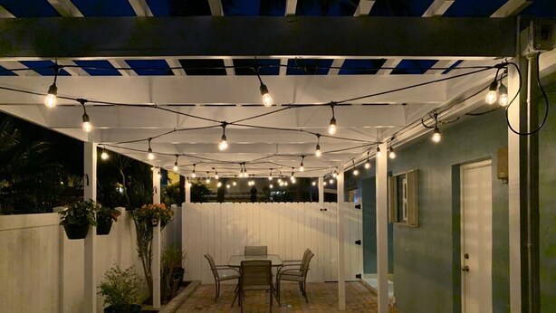 An outdoor patio area with a table and chairs, string lights overhead, and a white fence. - 2-Bedroom Apartment Just A Short Walk To Deerfield Beach Pier (Deerfield Beach)