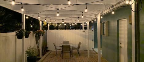 Een terras buiten met een tafel en stoelen, sfeerverlichting erboven en een witte omheining.