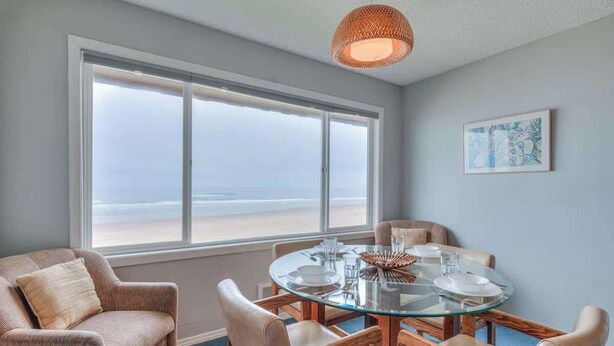 Ein Essbereich mit einem Glastisch, beigefarbenen Stühlen und Blick auf den Strand durch große Fenster.