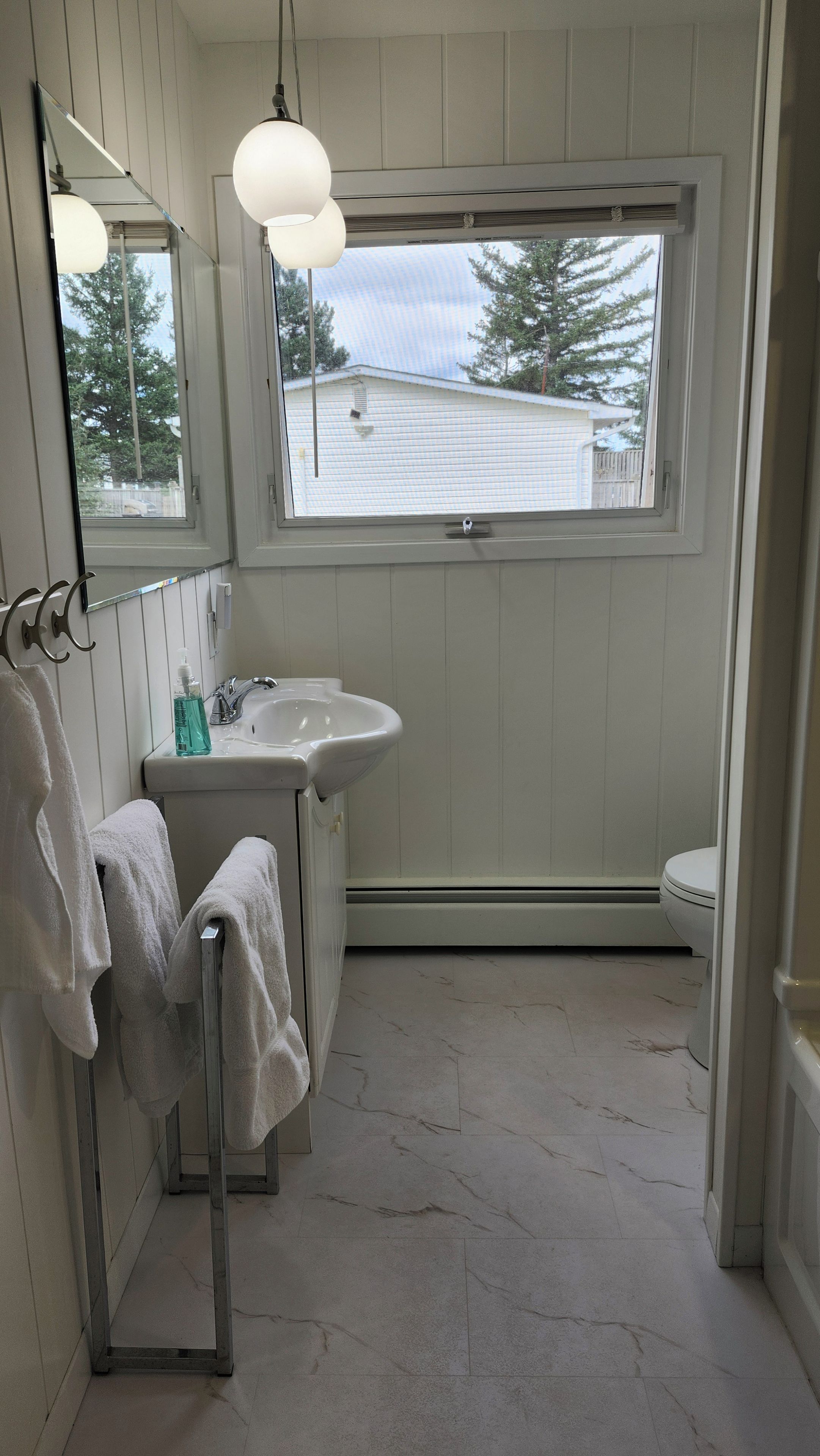 Ein Badezimmer mit einem weißen Waschbecken, einem Spiegel, einem Handtuchhalter und einem Fenster mit Blick auf ein Haus.