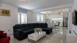 A modern living room with a sectional sofa, a glass coffee table, and a red armchair. A kitchen is visible in the background with white cabinets and stainless steel appliances. - Resort Villa San Lilin (Nerežišće)