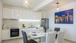 A modern kitchen with white cabinets, a built-in oven, and a dining area with a white table and chairs. - Resort Villa San Lilin (Nerežišće)