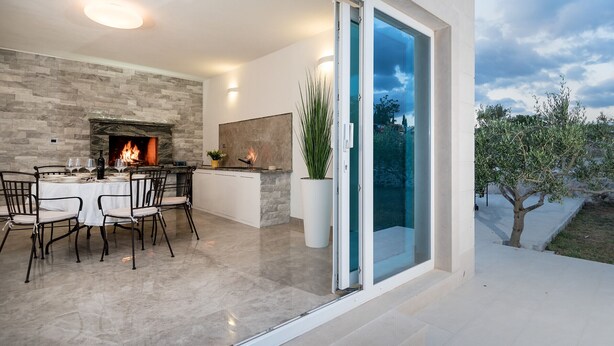 A modern dining area with a stone fireplace, a glass door leading to a balcony, and a view of the outdoors. - Resort Villa San Lilin (Nerežišće)