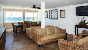 Un salon avec des meubles en osier, un coin repas avec une table et des chaises, une grande fenêtre offrant une belle vue et un téléviseur à écran plat fixé au mur.
