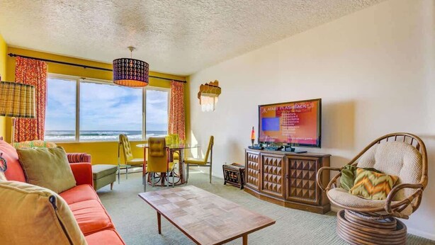 A living room with a sofa, a coffee table, a television, and a view of the ocean. - Retro Oceanfront Suite - Pool and Sauna - Second Floor - Sleeps 4 (Lincoln City)