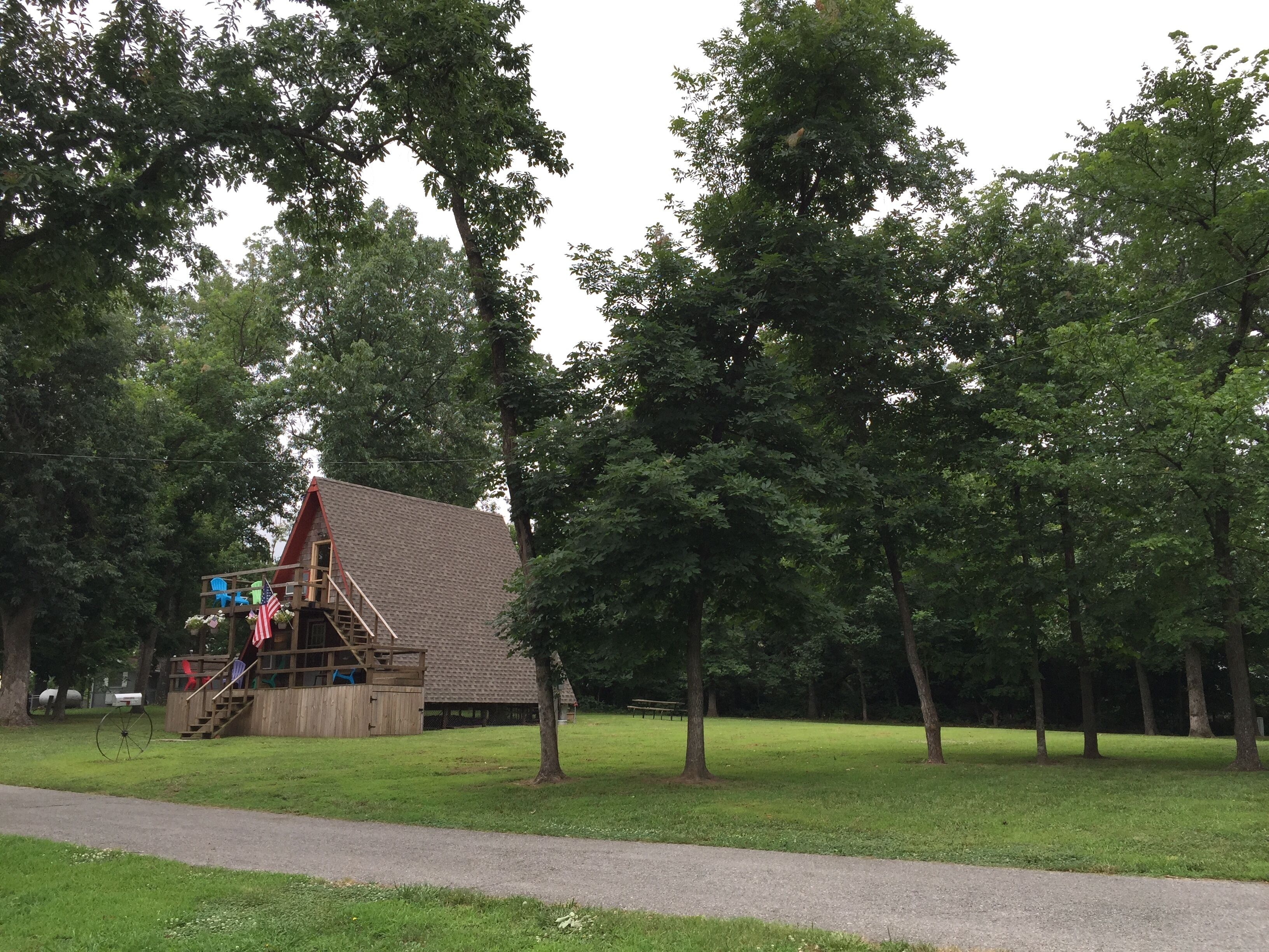 A rustic wooden cabin with a sloped roof, surrounded by lush green trees and a paved path.