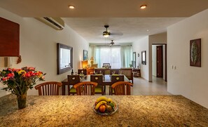 A dining area with a granite table, chairs, and a fruit bowl. There is a ceiling fan, a window with curtains, and a door in the background.