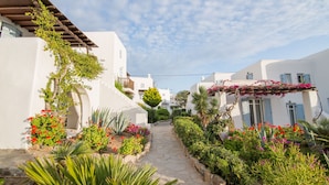 A narrow stone pathway flanked by white buildings with blue shutters, surrounded by lush greenery and flowering plants. - Pleiades Villa, Near Ornos Beach (Mykonos)