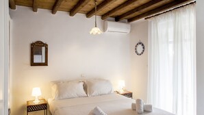 A bedroom with a bed, two bedside tables with lamps, a hanging light fixture, an air conditioning unit, a decorative mirror, and a window with sheer curtains. - Pleiades Villa, Near Ornos Beach (Mykonos)